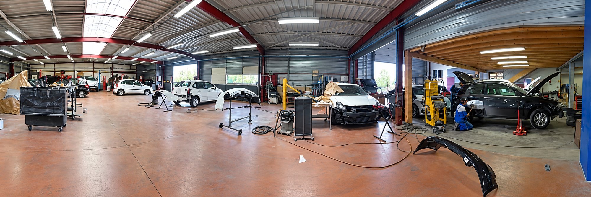 Vue panoramique d'un atelier de carrosserie où plusieurs voitures endommagées sont en cours de réparation et où un employé travaille près d'un véhicule noir.