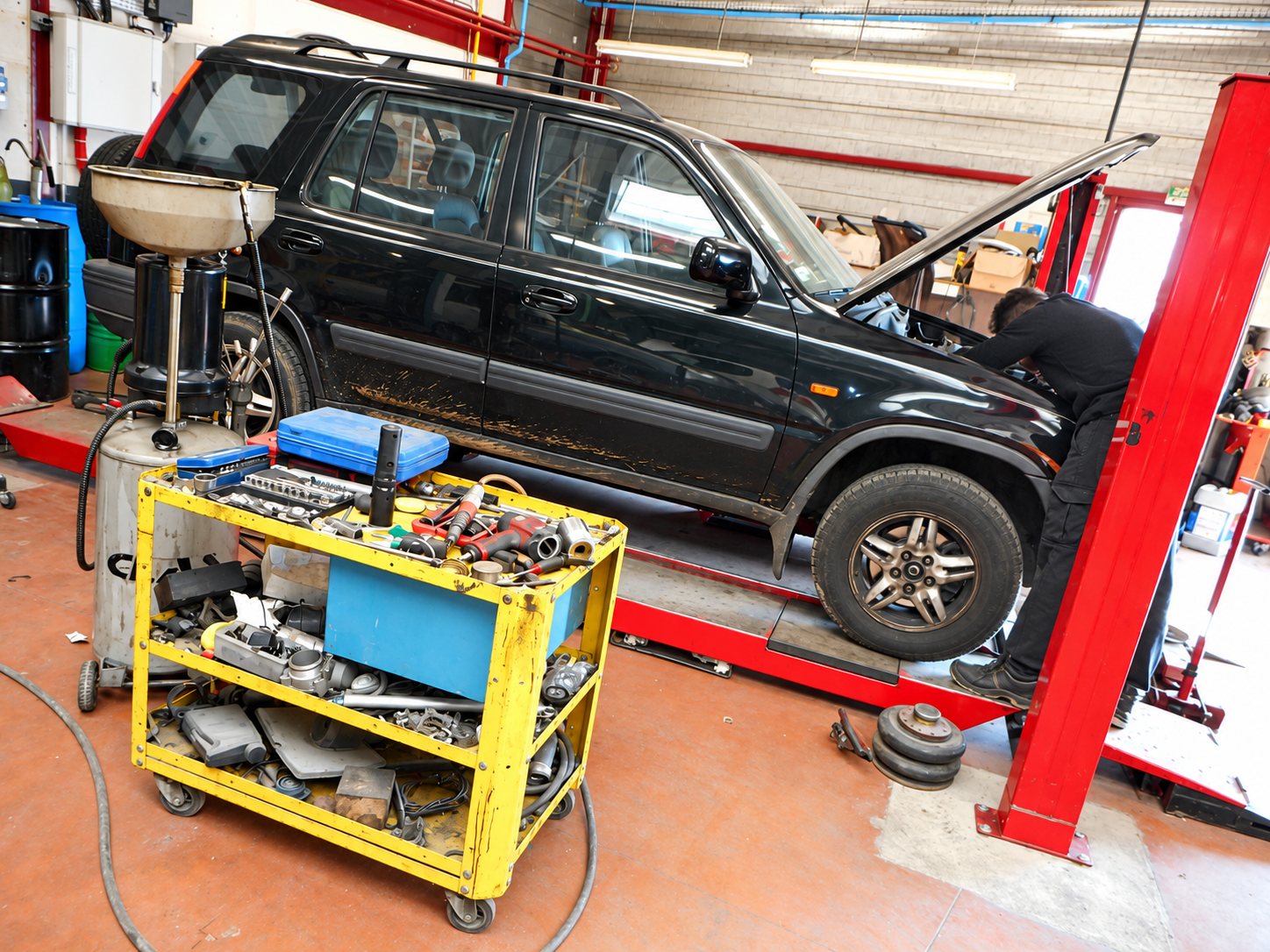 Dans un atelier de mécanique, un mécanicien penché sous le capot ouvert d'un SUV noir posé sur un pont élévateur rouge, à côté d'un chariot d'outils jaune et d'un récupérateur d'huile.