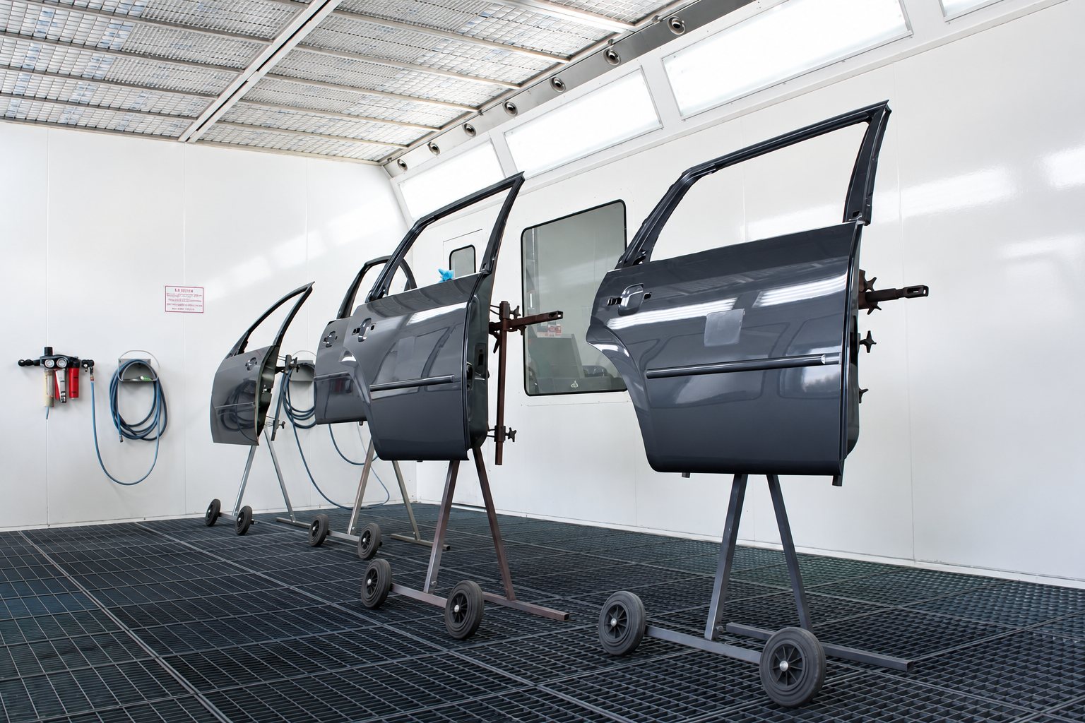 Quatre portières de voiture grises fixées sur des supports à roulettes dans une cabine de peinture blanche au sol grillagé.